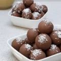 Two white bowls piled high with chocolate-covered Bounty balls sprinkled with shredded coconut, featuring a text overlay that reads "Bounty balls 3 ingredients