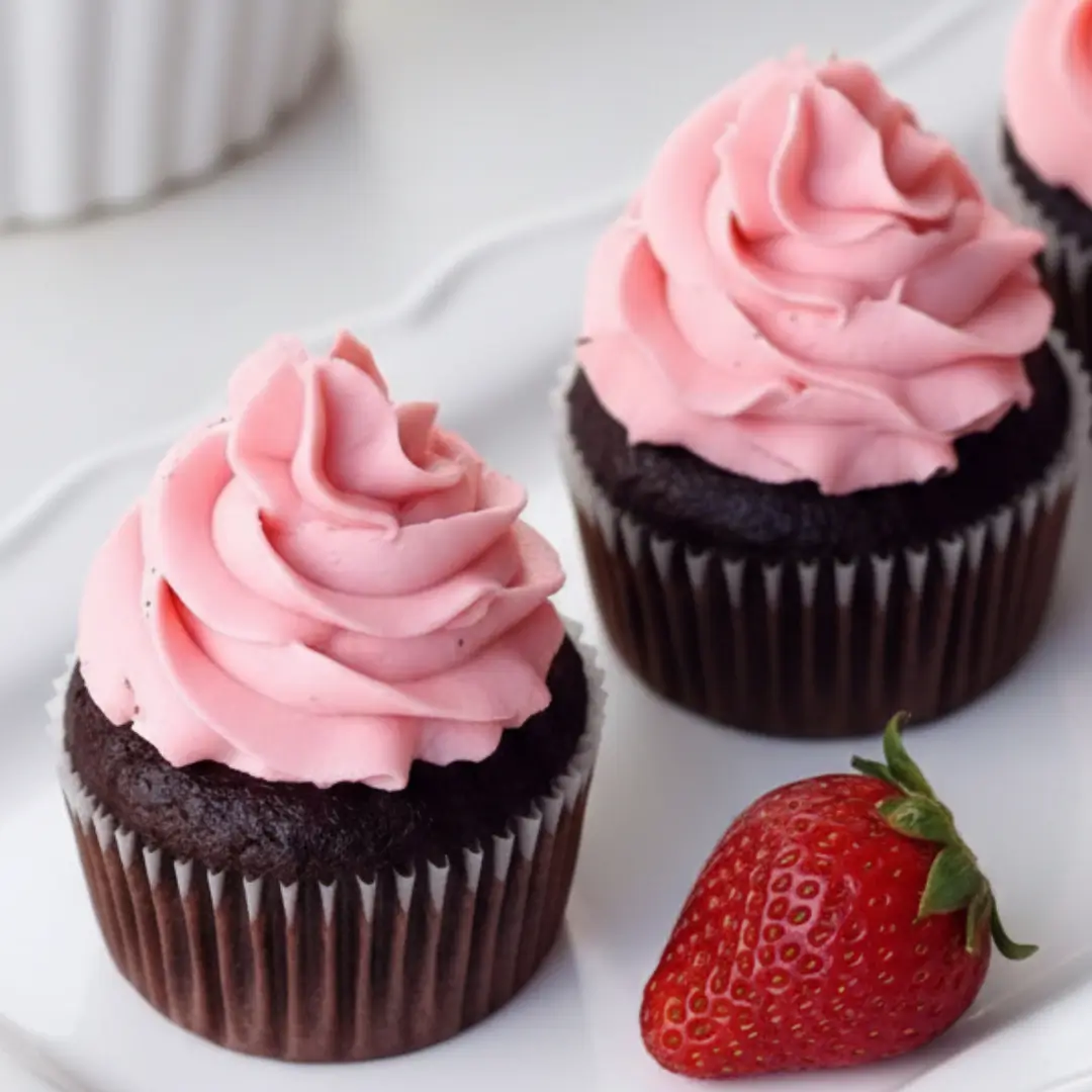 A close-up of decadent chocolate strawberry cupcakes featuring a rich chocolate base topped with a tall swirl of smooth pink frosting, sitting next to a fresh whole strawberry on a white platter.