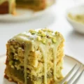 A close-up of a thick slice of pistachio coffee cake topped with a dripping green glaze and chopped pistachios, resting on a small plate with a fork next to it.pistachio coffee cake recipe