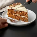 Cutting a slice of layered homemade carrot cake with thick cream cheese frosting and chopped pecans on a white dessert plate. Best Keto carrot cake