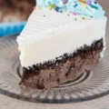 A slice of homemade Carvel ice cream cake featuring chocolate and vanilla layers separated by a crunchy fudge center, topped with blue frosting and rainbow sprinkles.