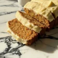 Close up of sliced Cinnamon Roll Carrot Cake Bread topped with thick, swirled cream cheese frosting on a white marble countertop.