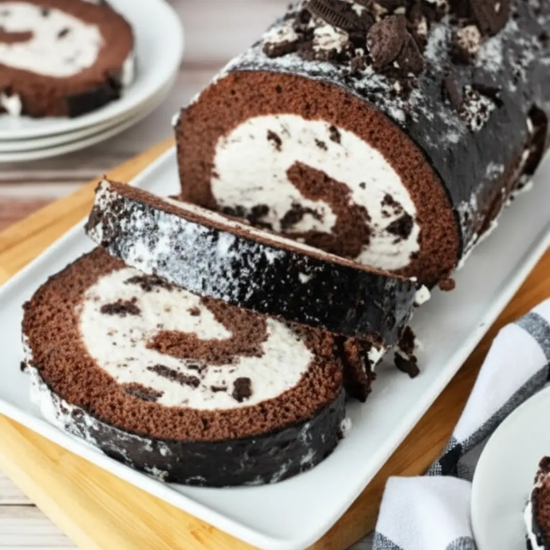 A partially sliced Homemade Oreo Ice Cream Cake Roll filled with creamy frosting and topped with crushed cookies on a white serving platter.