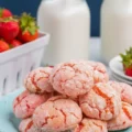 A stack of freshly baked, pink strawberry cake cookies with a powdered sugar crinkle finish, sitting on a light blue plate next to a basket of fresh strawberries.
