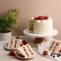 A sliced three-layer vanilla cake revealing a vibrant mixed berry fruit cake filling, displayed on a wooden cake stand with individual slices on plates.