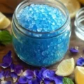 A close-up shot of a glass jar filled with bright blue coarse salt crystals, surrounded by scattered purple flower petals, lemon wedges, and fresh mint leaves on a wooden table. blue salt trick recipe