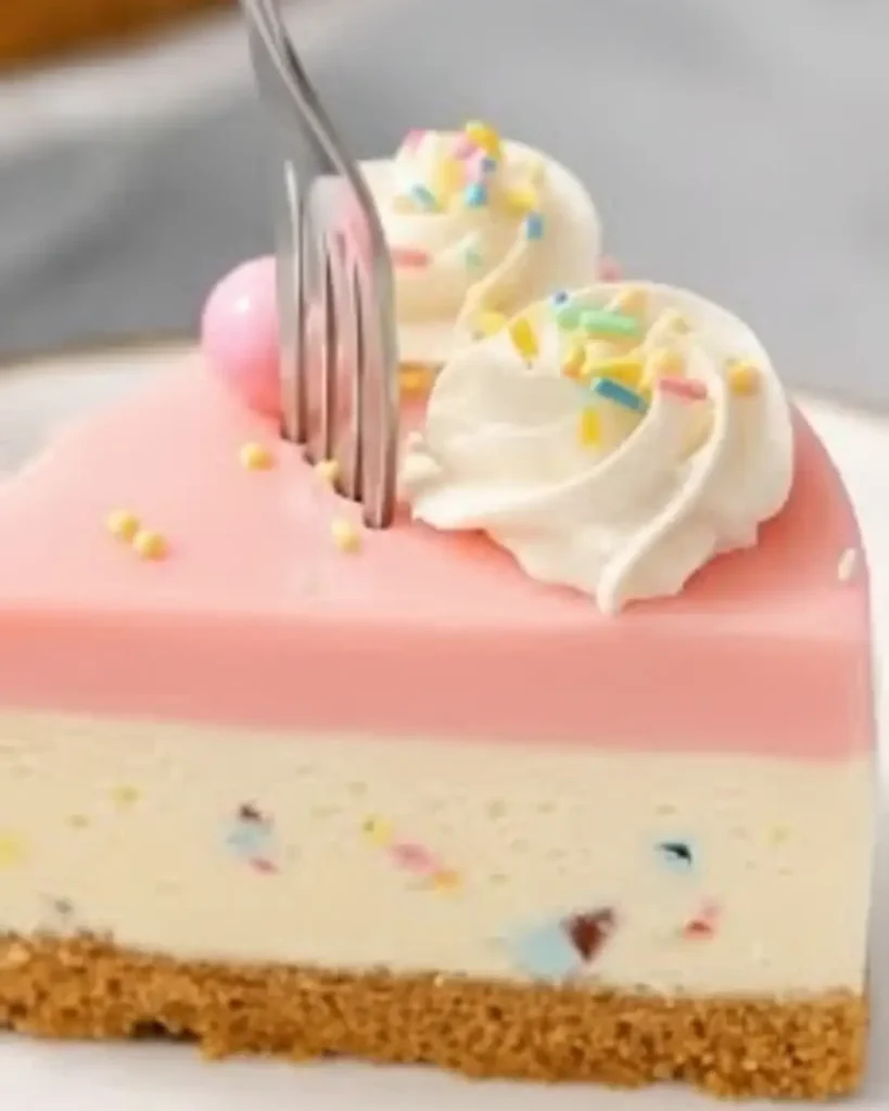 A close-up of a fork cutting into a slice of no bake Easter cheesecake with a graham cracker crust, a sprinkle-filled center, a smooth pink top layer, and whipped cream.