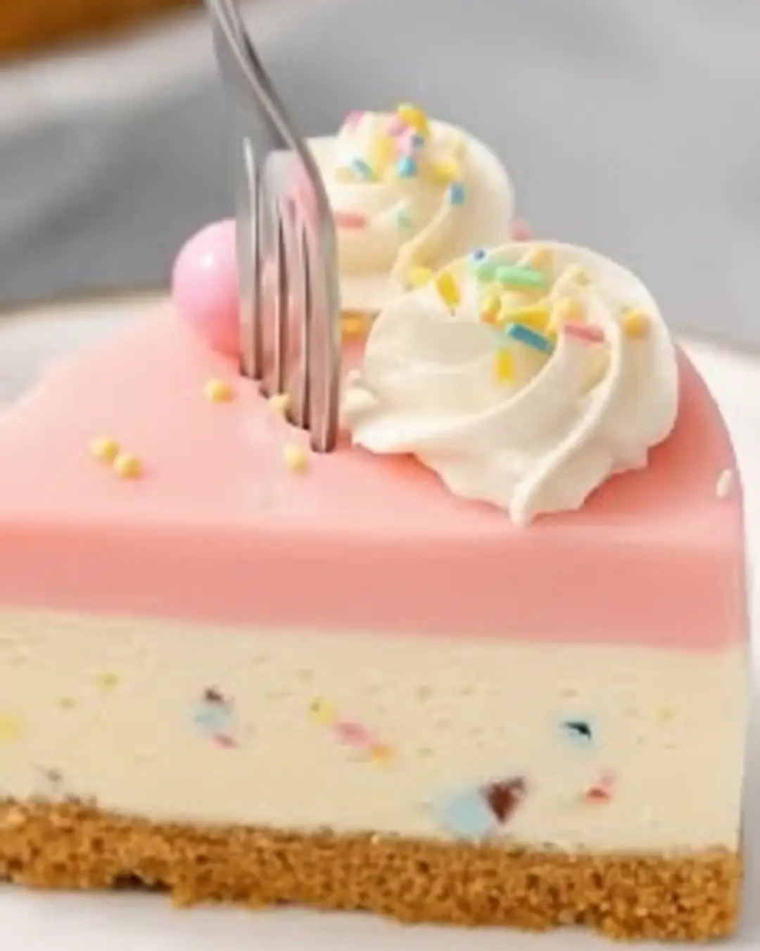 A close-up of a fork cutting into a slice of no bake Easter cheesecake with a graham cracker crust, a sprinkle-filled center, a smooth pink top layer, and whipped cream.