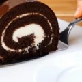 A close-up of a slice of chocolate sponge roll with a white cream filling and a glossy chocolate glaze being lifted by a metal cake server.