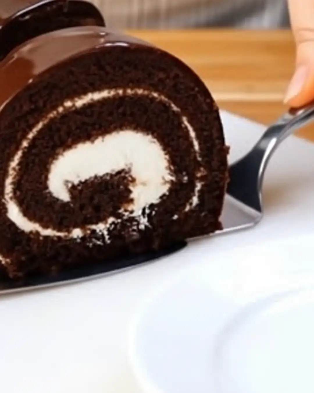A close-up of a slice of chocolate sponge roll with a white cream filling and a glossy chocolate glaze being lifted by a metal cake server.
