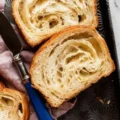 Two slices of a freshly baked sourdough croissant loaf revealing a flaky, airy crumb, resting on a dark patterned tray next to a blue-handled knife.
