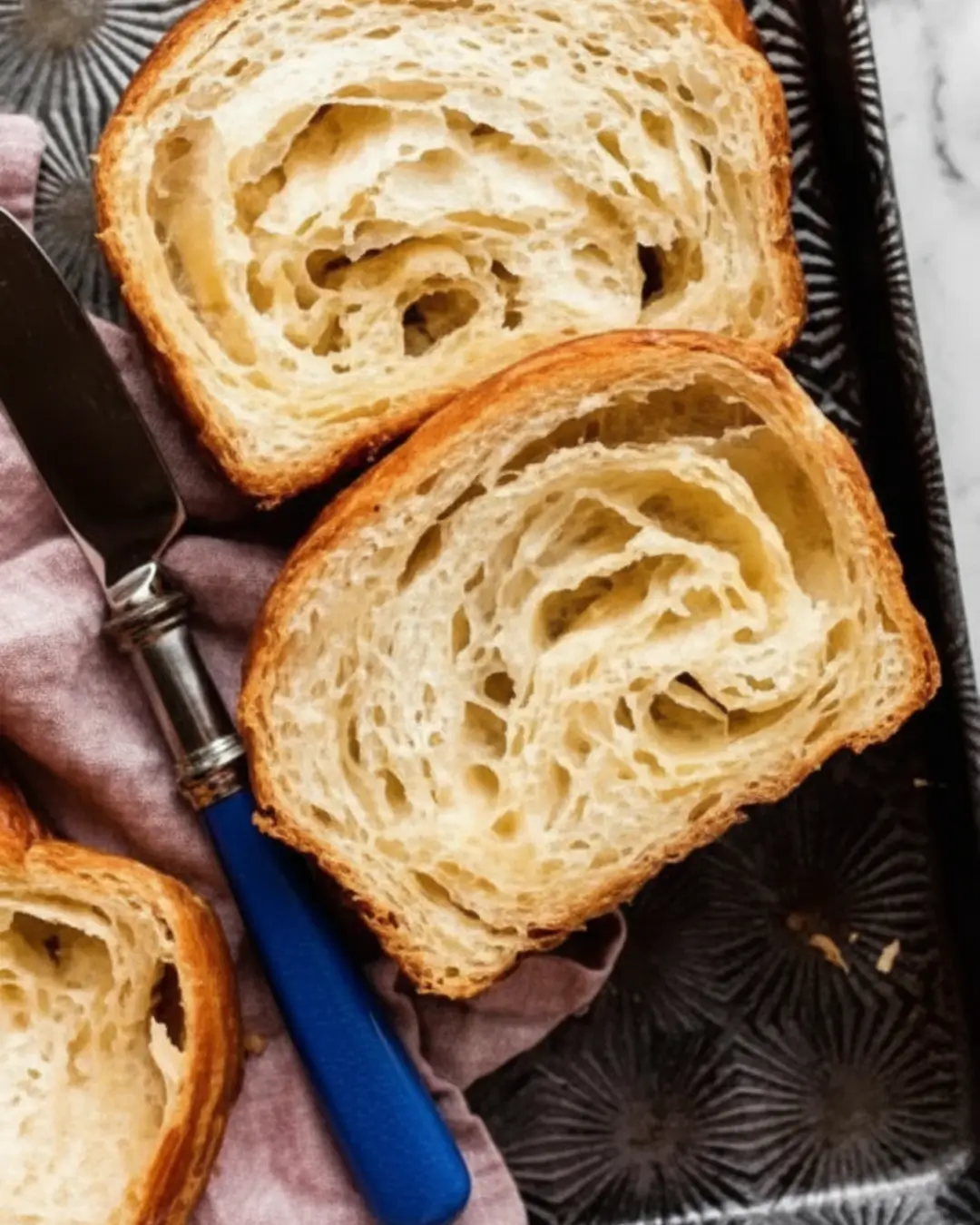 Two slices of a freshly baked sourdough croissant loaf revealing a flaky, airy crumb, resting on a dark patterned tray next to a blue-handled knife.