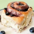 A close-up of a freshly baked, fluffy pastry from a sourdough blueberry rolls recipe, topped with white icing glaze and surrounded by fresh blueberries.