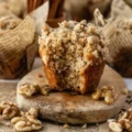 A partially eaten banana bread muffin with a crumbly walnut streusel topping on a rustic wooden board, surrounded by loose walnuts and cinnamon sticks.