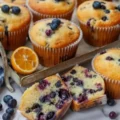 A close-up of freshly baked lemon blueberry muffins on a rustic metal tray, with one muffin cut in half to show the moist crumb and juicy blueberries inside, garnished with fresh berries and a slice of lemon.