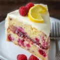 A slice of layered raspberry lemon cake on a white plate, frosted with white icing and garnished with fresh raspberries and a lemon twist.