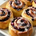 Close-up view of warm, golden-brown Huckleberry Cinnamon Rolls filled with dark berries and generously drizzled with white icing on a white serving platter.