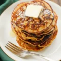 A close-up of a freshly cooked stack of homemade banana pancakes topped with melting butter and syrup on a white plate with a silver fork.