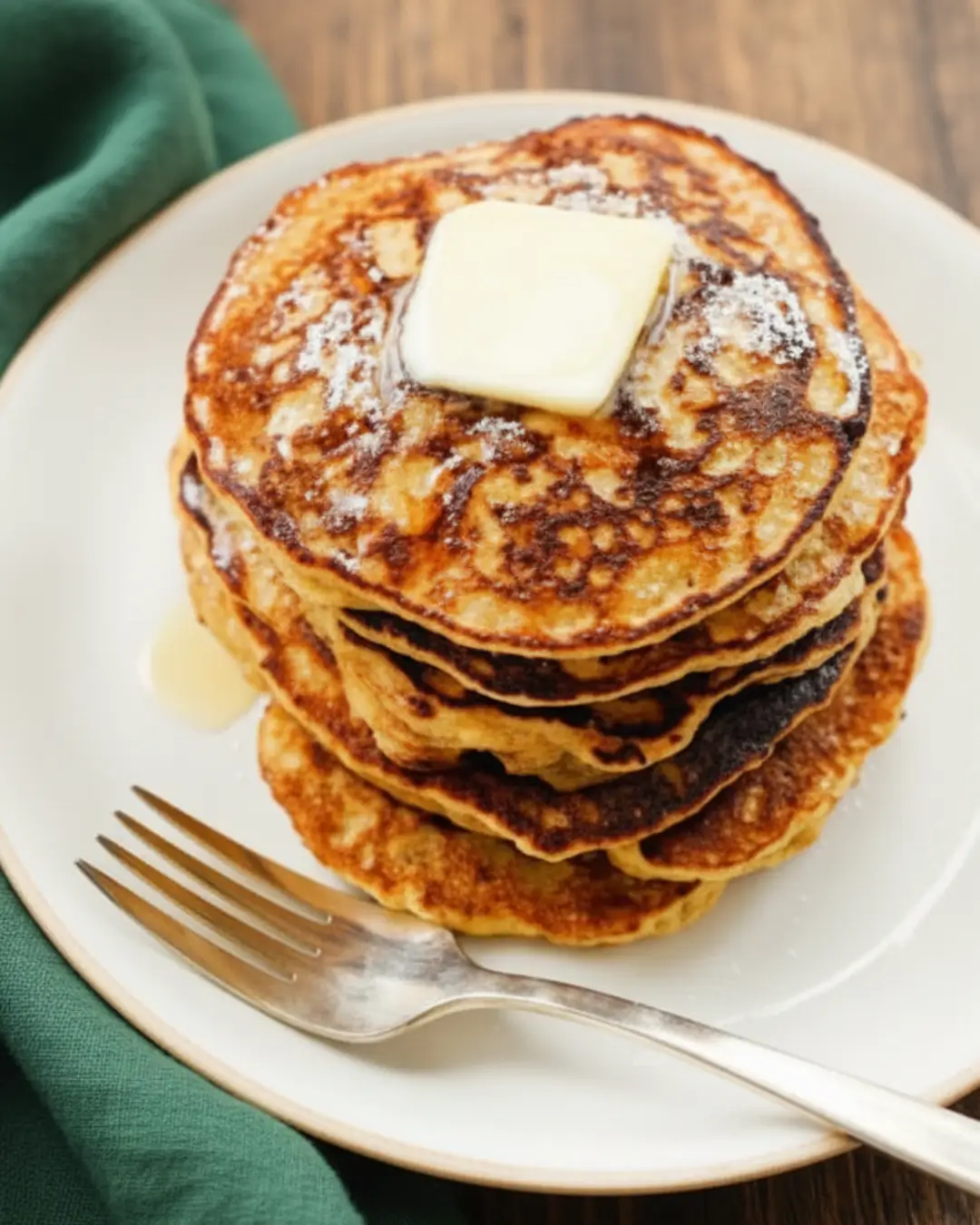 A close-up of a freshly cooked stack of homemade banana pancakes topped with melting butter and syrup on a white plate with a silver fork.