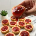 A hand holding a mini strawberry cheesecake with sweet strawberry topping dripping down onto a white plate filled with more mini cheesecakes.