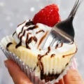 Close-up of a fork cutting into a decadent Brownies Bottom Mini Cheesecake garnished with whipped cream, chocolate drizzle, and a fresh raspberry.