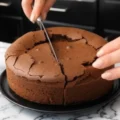 Hands slicing a decadent, dense flourless chocolate cake resting on a black serving plate over a white marble countertop.