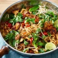 A skillet filled with a savory noodle stir-fry recipe featuring glazed chicken, snow peas, bean sprouts, red chili peppers, fresh cilantro, and lime wedges.
