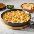 A cast iron skillet filled with hot baked buffalo chicken dip topped with blue cheese crumbles, hot sauce, and green onions.