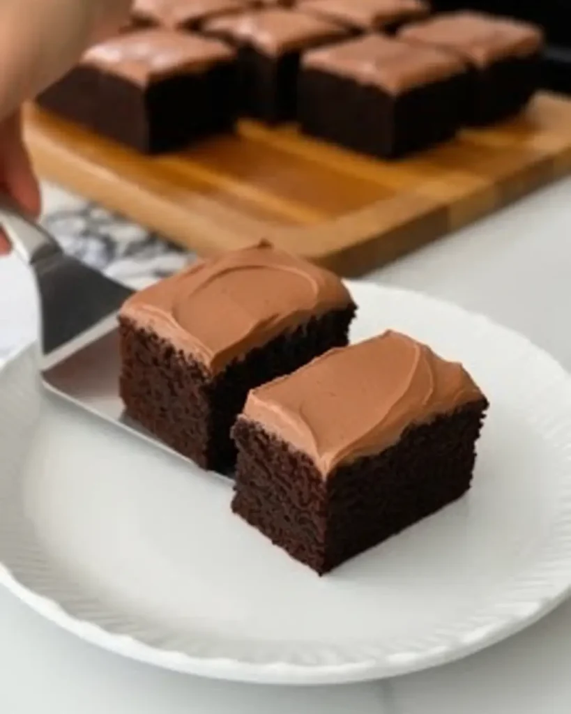 A serving spatula placing a frosted slice of the best chocolate cake recipe onto a white plate next to another slice.
