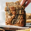 Hand picking up the top slice from a tall stack of freshly baked gluten-free banana bread topped with caramelized banana strips.