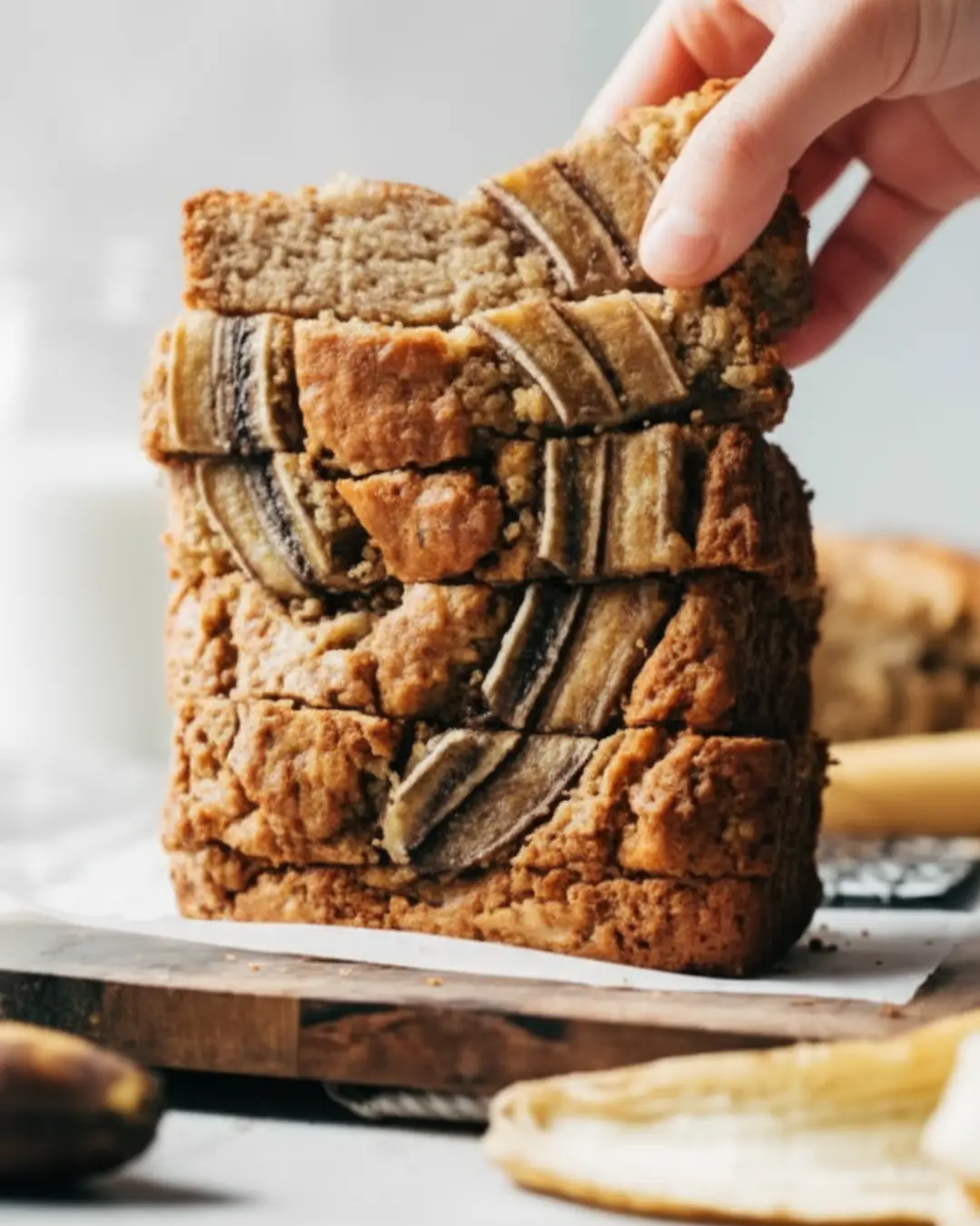 Hand picking up the top slice from a tall stack of freshly baked gluten-free banana bread topped with caramelized banana strips.
