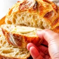 A hand holding a buttered slice of easy homemade crusty artisan bread next to the freshly baked, sliced loaf.