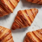Top-down view of several golden-brown, flaky pastries, highlighting the crispy layers of freshly baked croissant bread resting on a white marble countertop.