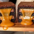 Close-up cross-section of a 6-layer Caramel Brownie cut in half, revealing thick, fudgy chocolate layers, embedded chocolate bars, and warm, gooey caramel oozing out onto a wooden board.