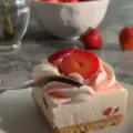 A close-up of a spoon cutting into a creamy slice of Strawberries and Cream No-Bake Cheesecake Bars topped with whipped cream and fresh strawberries.