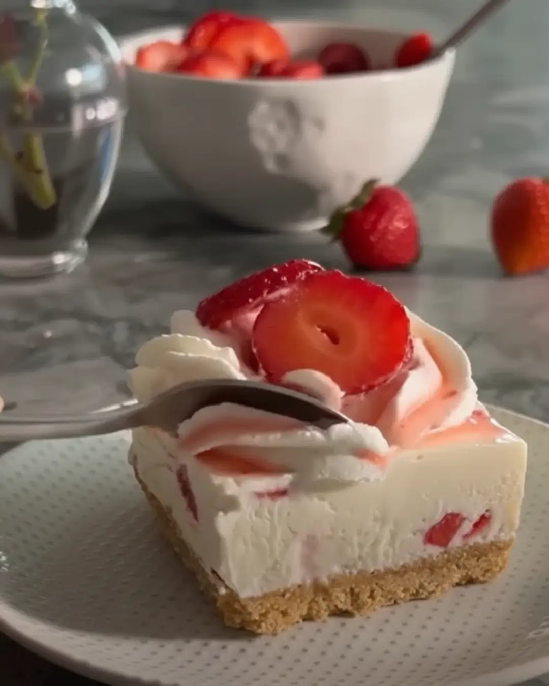 A close-up of a spoon cutting into a creamy slice of Strawberries and Cream No-Bake Cheesecake Bars topped with whipped cream and fresh strawberries.