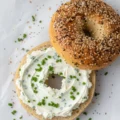Overhead view of a freshly made everything bagel recipe spread with chive cream cheese, surrounded by fresh chives and seasoning.