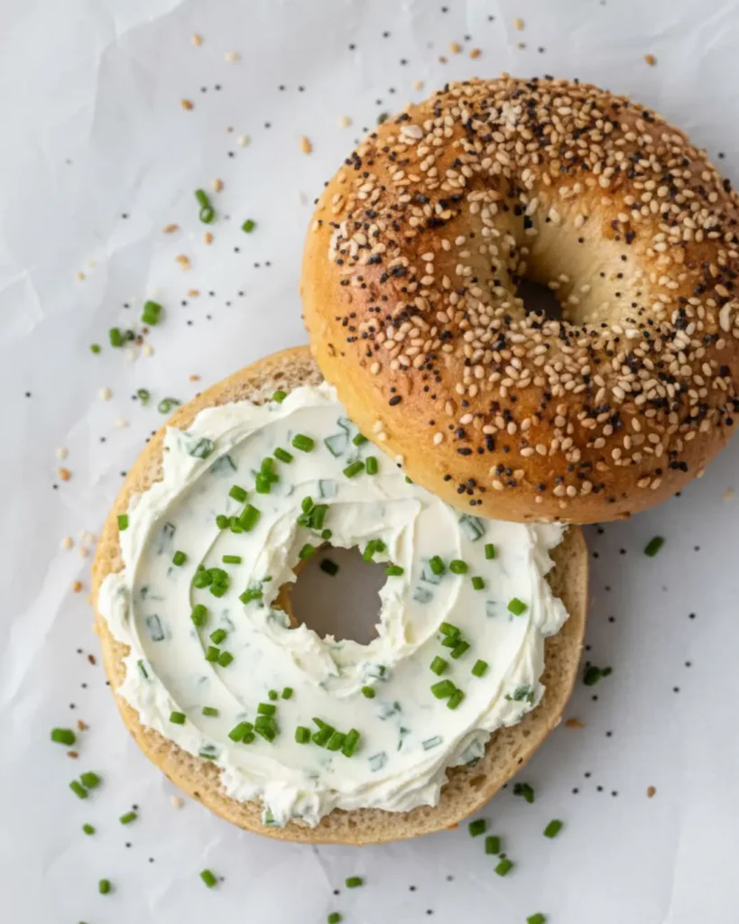 Overhead view of a freshly made everything bagel recipe spread with chive cream cheese, surrounded by fresh chives and seasoning.