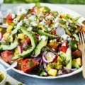 A large white bowl filled with a colorful chopped summer salad containing red and yellow cherry tomatoes, diced cucumber, sweet corn, sunflower seeds, and crumbled feta cheese.