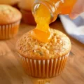 Hand pouring a thick orange glaze from a glass jar onto a freshly baked Best Ever Banana Muffin.