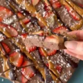 A round, uncut chocolate frozen yogurt bark topped with sliced strawberries, peanut butter, chocolate drizzle, and coconut flakes on a blue plate.