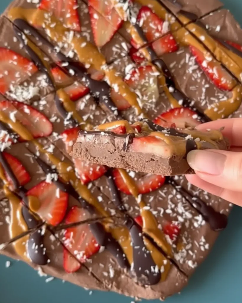 A round, uncut chocolate frozen yogurt bark topped with sliced strawberries, peanut butter, chocolate drizzle, and coconut flakes on a blue plate.