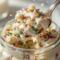 A silver spoon lifting a rich bite of colorful homemade dot cake and white vanilla frosting out of a clear glass jar, with loose sprinkles falling through the air.